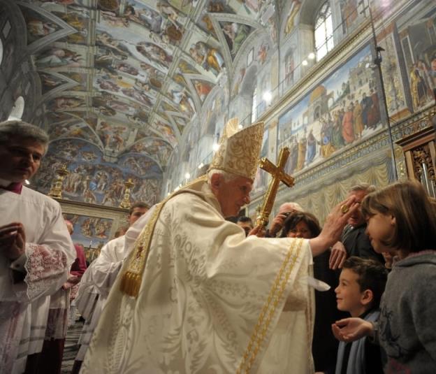 A man in cream robes with a tall hat and carrying a large gold cross reaches out his right hand toward the forehead of a child in a grey sweater, while others watch. Paintings can be seen all over the walls and ceiling of the room.