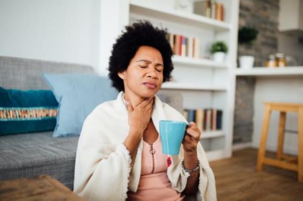 woman, fallen ill is staying at home wrapped in a blanket socially distancing and quarantining herself, feeling her throat hurt and being sore, havin<em></em>g a cup of hot tea