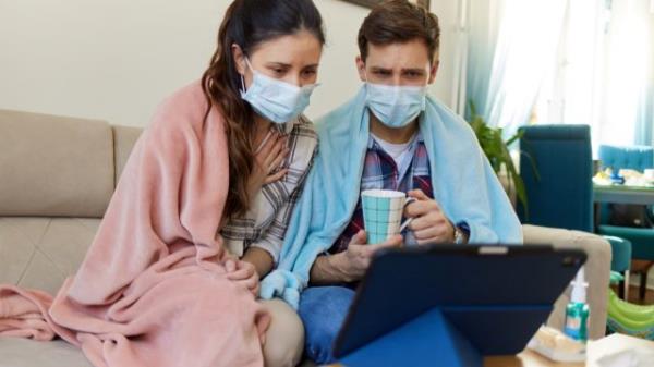 Scared couple in masks sitting on sofa and co<em></em>nsulting o<em></em>nline with a doctor over digital tablet