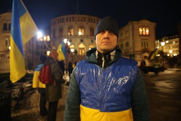 A man dressed in the Ukrainian flag stands in front of the Norwegian parliament building in the dark during a protest.