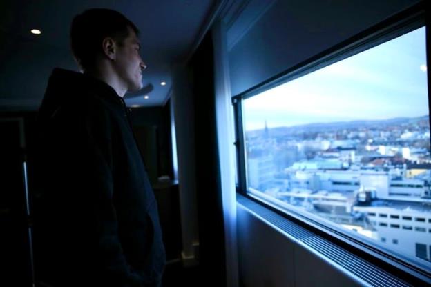 A man in a black hoodie looks out a window, with a city landscape spread out below.