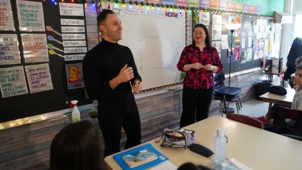A man wearing a black shirt and pants speaks next to a classroom whiteboard, as a woman in a red-and-black patterned shirt next to him smiles.   