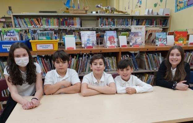 Five elementary school students sit together at a table in a school library, with many bookshelves filled with books seen behind them.