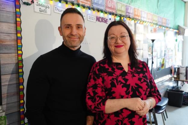 A man in a black turtleneck and woman in a red-and-black patterned shirt smile as they stand next to a whiteboard, with brightly coloured classroom displays behind them.